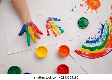 Little child making hand print on paper with painted palm at white table indoors, top view