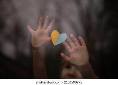 Little Child, An Orphan, Holding A Heart Shaped Paper In Ukrainian Flag Colors Of Yellow And Blue Against A Window.