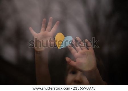 Little child, an orphan, holding a heart shaped paper in Ukrainian flag colors of yellow and blue against a window.