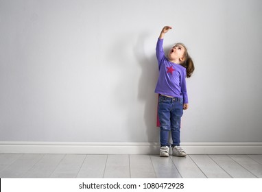 Little Child Is Playing Superhero. Kid Is Measuring The Growth On The Background Of Wall. Girl Power Concept. 