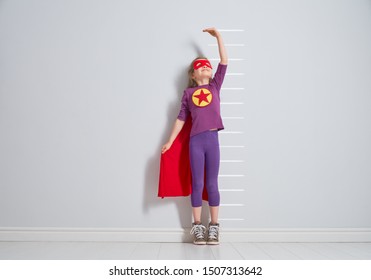 Little Child Is Playing Superhero. Kid Is Measuring The Growth On The Background Of Wall. Girl Power Concept. 