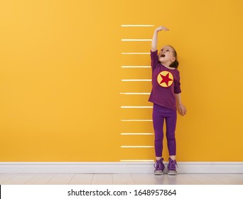 Little Child Is Playing Superhero. Kid Is Measuring The Growth On The Background Of Bright Color Wall. Girl Power Concept. Yellow, Red And Violet Colors.                               