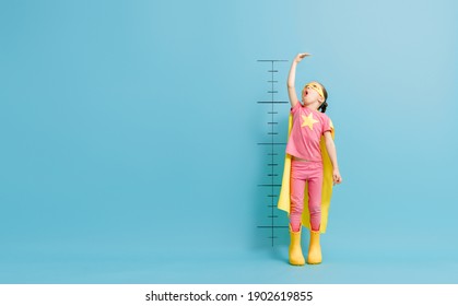 Little Child Is Playing Superhero. Kid Is Measuring The Growth On The Background Of Color Wall. Girl Power Concept. 