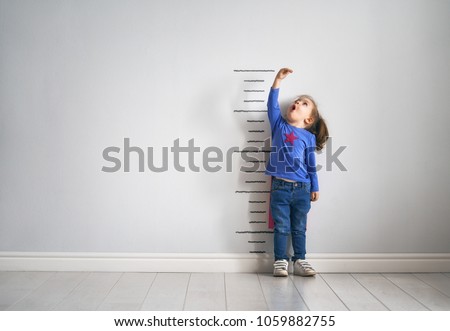 Little child is playing superhero. Kid is measuring the growth on the background of wall. Girl power concept. 