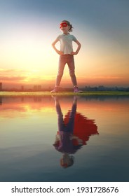 Little Child Playing Superhero And Looking At Her Reflection In Water Surface. Kid On The Background Of Sunset Sky. Girl Power Concept