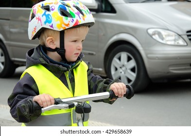 Little Child Rides A Bike In The City Traffic. There Is A Car Driving In The Background. Boy Is Marked By Yellow Reflective Vest And Helmet Because Of Visibility And Safety.