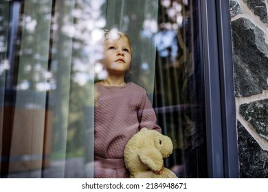 Little child standing alone with teddy bear behind the window, photo trough glass. - Shutterstock ID 2184948671