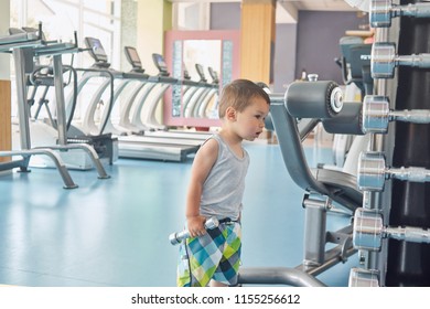 Little Child Standing In Gym In Front Of Metallic Dumbbells And Racetracks. Searching For Father, Looking Dissapointed, Interested, Maintaining Healthy Lifestyle, Cute Face, Big Eyes, Plump Lips.