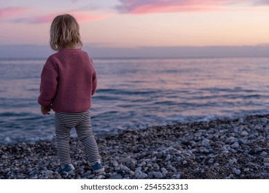 A little child stands on a pebble beach, admiring the vibrant sunset reflections on the water. The tranquil scene captures the beauty of nature as dusk approaches. Blonde toddler in pink sweater - Powered by Shutterstock