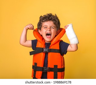 Little Child Superhero Raises Arms And Shows Muscles. Strong Confident Boy Shouts Loudly. Kid With Broken Arm In Orange Inflated Lifejacket Isolated On Yellow. Swimming Aid For Kids. Little Hero.