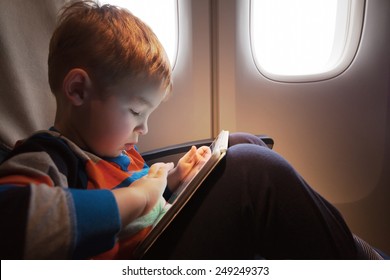 Little Child With Tablet Computer On The Lap Sitting By The Illuminator In The Plane