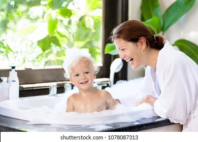 Little Child Taking Bubble Bath In Beautiful Bathroom With Big Garden View Window. Mother Washing Baby. Kids Hygiene. Shampoo, Hair Treatment And Soap Foam For Children. Mom Bathing Kid In Large Tub. 