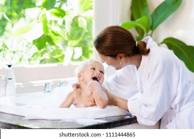 Little Child Taking Bubble Bath In Beautiful Bathroom With Big Garden View Window. Mother Washing Baby. Kids Hygiene. Shampoo, Hair Treatment And Soap Foam For Children. Mom Bathing Kid In Large Tub. 