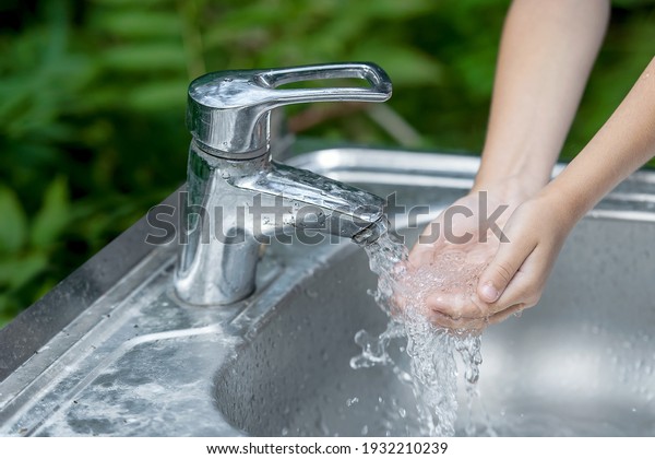 Little child washing hands with clean running\
water in the park. Coronavirus prevention hand hygiene. Concept\
hygiene, clean and health care. Flowing water from tap. Outdoor.\
Natural green background