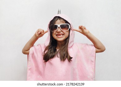 Little crazy happy girl in glasses and in a pink poncho towel shows her teeth and closes her ears on a white background, childish behavior, light, day, space