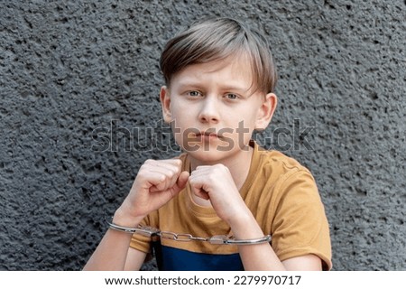 A little criminal is handcuffed against a gray wall, an emotional portrait. Concept: juvenile delinquency, petty theft and theft, incarceration.