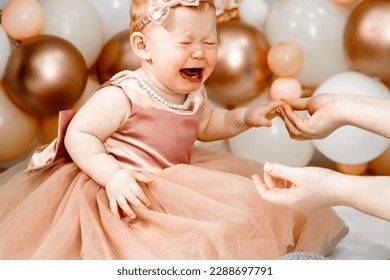 Little crying unhappy redhead baby girl celebrates first birthday anniversary. 1 year family party Professional photoshoot in photo studio. Cute adorable red hair kid in pink dress.Home children room.