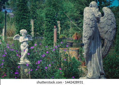 Little Cupid Angel Statue Standing In The Garden Amid Purple Flowers