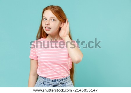 Little curious nosy redhead kid girl 12-13 years old wearing pink striped t-shirt try to hear you overhear listening intently isolated on pastel blue background studio. Lifestyle childhood concept.