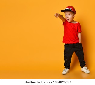 Little cute 4 year old boy in wide dark trousers with pockets and a red T-shirt. Funny hat with shark teeth. Standing on a yellow background shows a finger at something interesting.