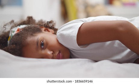 Little Cute African American Girl Lying On Bed And Smiling At Camera. Side View Of Pretty Afro Child Relaxing On Bed And Looking At Camera Playing At Home. Childhood Concept