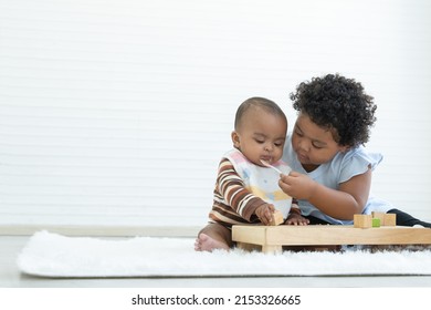 Little Cute African Older Sister Try To Feeding Her Newborn Sister Food While Baby Playing Toys Sitting On Floor At Home. Relationship Of Siblings Concept. White Background. Copy Space