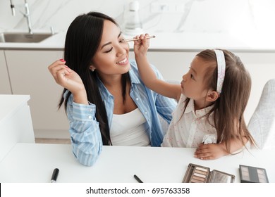 Little Cute Asian Girl Applying Make Up On Her Mothers Face While Sitting At The Table At Home