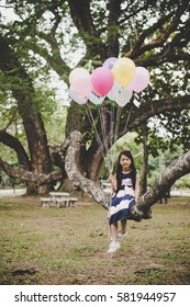 Little cute asian girl sitting on tree branch with colorful of balloons in hand, Relaxing enjoy holiday.
