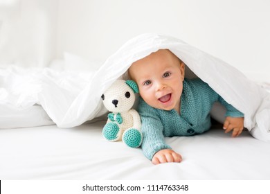 Little Cute Baby Boy, Child In Knitted Sweater, Holding Knitted Toy, Smiling Happily At Camera In White Sunny, Bright Bedroom