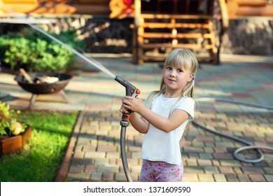 Little Cute Baby Girl Watering Fresh Green Grass Lawn Mear House Backyard On Bright Summer Day. Child Having Fun Playing With Water Hose Sprinkler