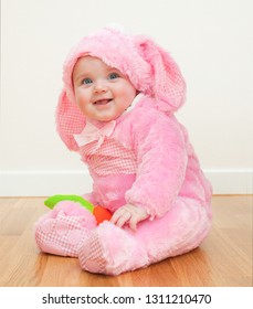 Little Cute Baby In A Pink Bunny Suit. The Child Sits On The Wooden Floor. 