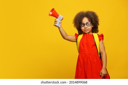 Little Cute Black Girl In Red Dress Holding In Hand And Speaking In Electronic Gray Megaphone On Yellow Background. Kids Childhood Lifestyle Concept. Copy Space