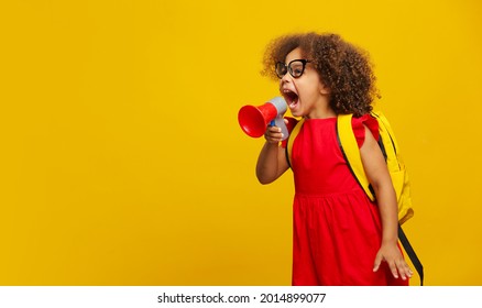 Little Cute Black Girl In Red Dress Holding In Hand And Speaking In Electronic Gray Megaphone On Yellow Background. Kids Childhood Lifestyle Concept. Copy Space