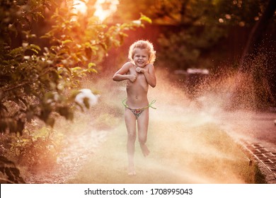 Little cute blond girl in bikini running on the grass under splashing water streams in sunset