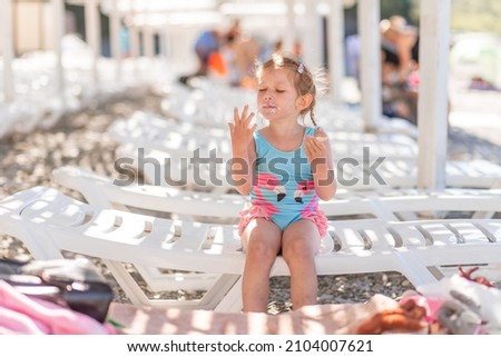 
A little cute blonde girl in a swimsuit sits on a sun lounger on the beach and licks her fingers in ice cream. The girl is eating ice cream. Summer rest.