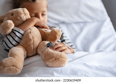 Little cute boy hugging teddy bear with intravenous drip in hand, sleeping under bed sheet in hospital. Sick child with pulse oximeter on finger resting with his toy on gurney at pediatric ward. - Powered by Shutterstock