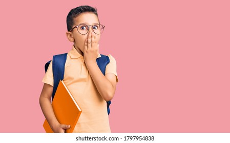 Little cute boy kid wearing school bag and holding book covering mouth with hand, shocked and afraid for mistake. surprised expression 