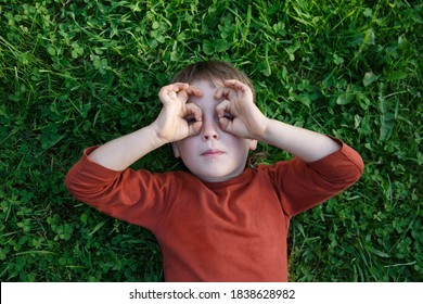 A little cute boy is lying on the green grass depicting glasses with his hands. - Shutterstock ID 1838628982
