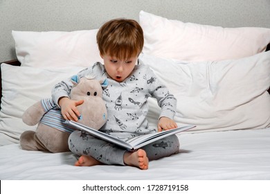 Little cute boy in pajamas reading a toy book sitting on the bed. Bedtime stories before bed.