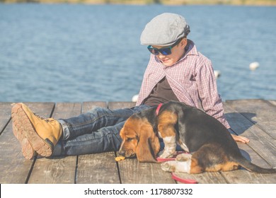 Little Cute Boy Is Sitting By The River With His Dog. They Enjoy Together On A Beautiful Sunny Day. Child Playing With His Puppy. Growing Up, Love For Animals - Dogs, Free Time, Travel, Vacation.