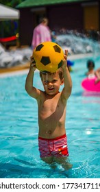 Little Cute Boy Standing Knee-deep In Clear Water Holding Yellow Ball For Football Over Head In Water Park