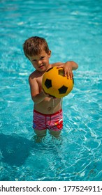 Little Cute Boy Standing Knee-deep In Clear Water Holding Yellow Ball For Football In A Water Park