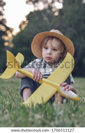 little cute boy in a straw hat sits on the grass with a big yellow plane. summer. sunset