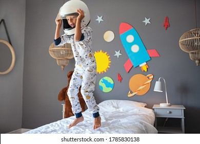 Little Cute Boy Wearing Cosmonaut Costume And Jumping On Bed With Rocket And Planets On Wall. Child Wearing Astronaut Helmet Playing And Dreaming Of Becoming A Spaceman. Joyful Kid Having Fun.