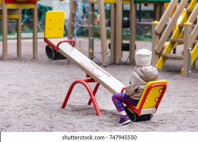 Little Cute Caucasian Girl In Jacket And Hat Sitting Alone On A Seesaw Swing At A Playground Outdoors. Loneliness Mood Concept.