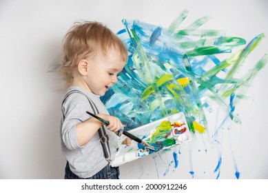 Little cute child and brush   palette paints in his hands draws white wall  Happy childhood 