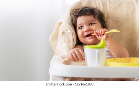 Little cute child celebrating her first birthday with cake at home. Baby adorable girl with apron holding spoon in her hand while sitting on the chair eating cake sloppy her face.