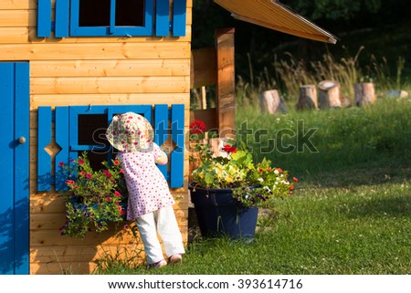 little cute curios girl looking into the blue window of wooden playhouse in the countryside garden with blooming flowers