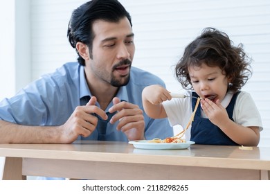 Little Cute Daughter Trying To Eat Spaghetti With Spoon By Herself At Home And Caucasian Young Father Looking And Cheer Her Up. Adorable Kid Girl Enjoy Eating With Face Is Mess Up With Ketchup