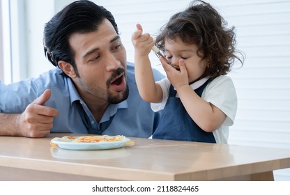 Little Cute Daughter Trying To Eat Spaghetti With Spoon By Herself At Home And Caucasian Young Father Looking And Cheer Her Up. Adorable Kid Girl Enjoy Eating With Face Is Mess Up With Ketchup